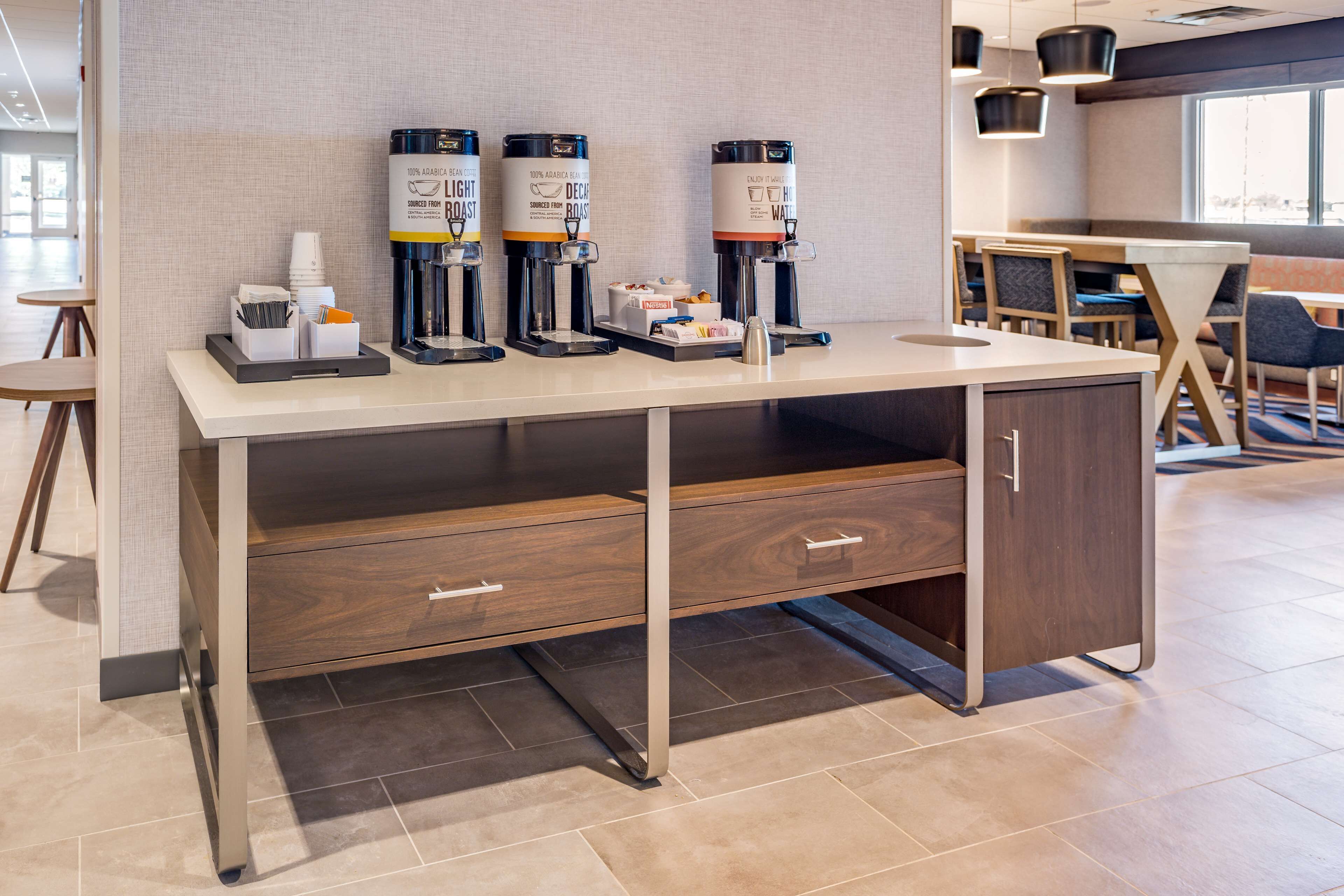 A modern hotel room with a long counter, three coffee machines, a small refrigerator, and a seating area with tables and chairs.