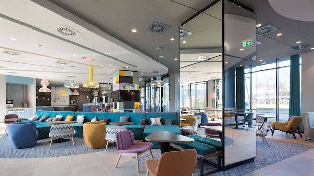 A modern lounge area with seating arrangements, a kitchen in the background, and large windows providing natural light.