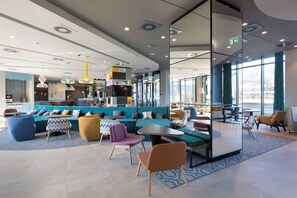 A modern lounge area with seating arrangements, a kitchen in the background, and large windows providing natural light. - Hampton by Hilton Krakow Airport (Zabierzow)