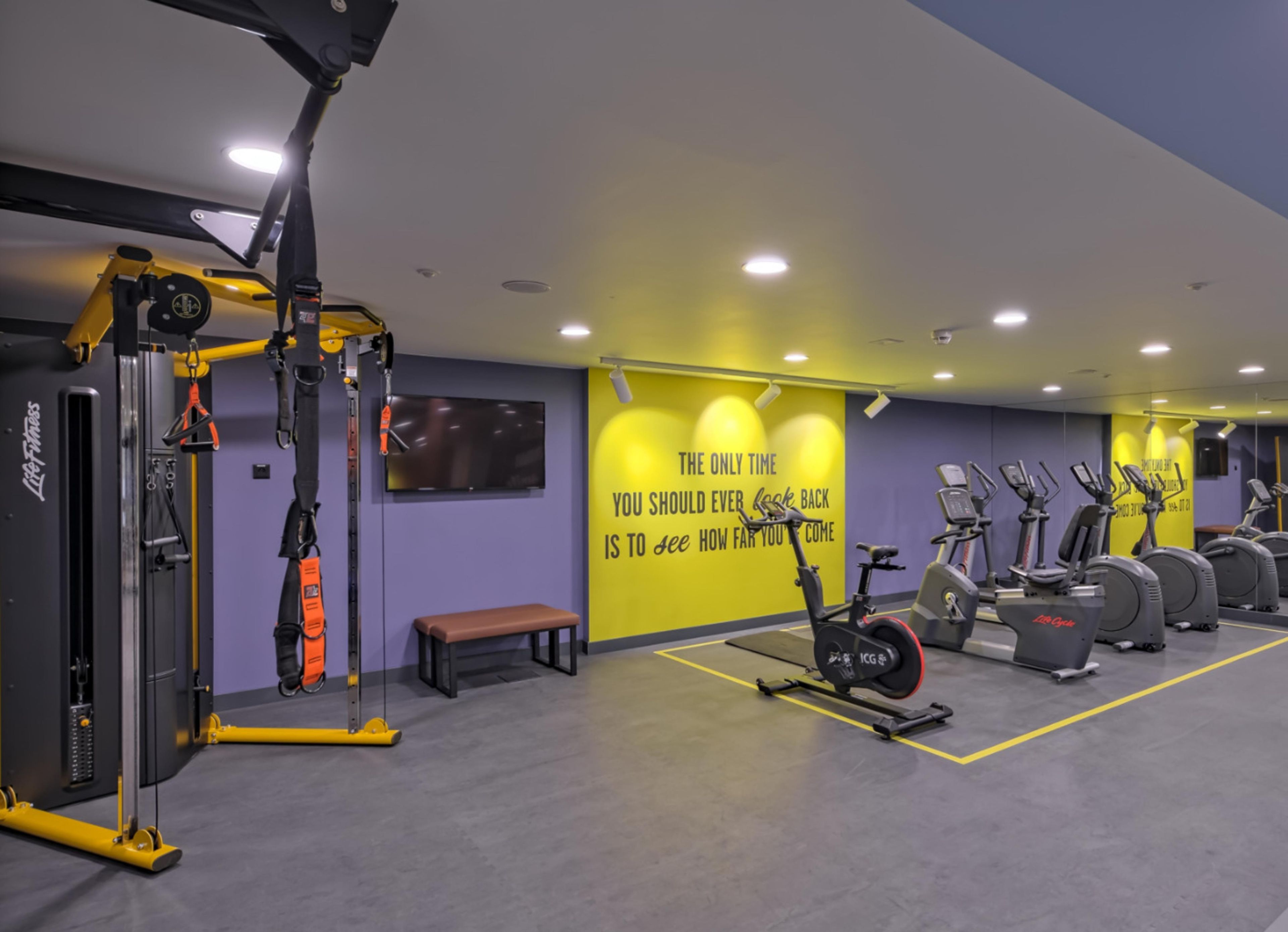 Een sportschool met trainingsapparatuur, een bankje en motiverende tekst op de muur.