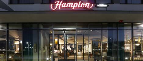 Una entrada de hotel moderna con fachada de vidrio y un letrero de neón que dice "Hampton by Hilton".