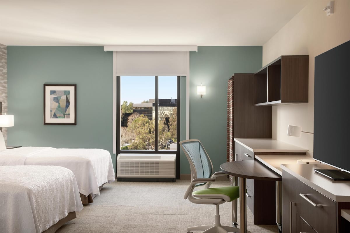 Habitación de hotel con dos camas, un escritorio con silla, una ventana con vistas a edificios y un televisor montado.