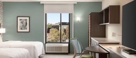 Habitación de hotel con dos camas, un escritorio con silla, una ventana con vistas a edificios y un televisor montado.