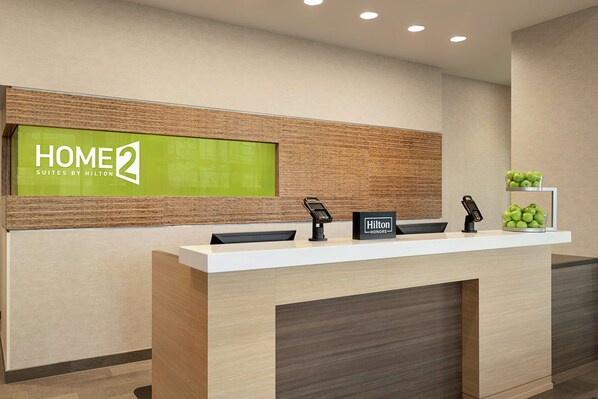 Un comptoir d’accueil d’hôtel avec une enseigne « HOME 2 Suites by Hilton », une plaque « Hilton Honors », un téléphone et une présentation de fruits.