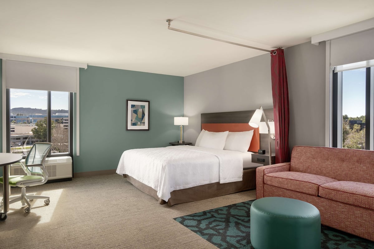 Habitación de hotel con cama, sofá, escritorio con silla, ventana con vistas y una lámpara.