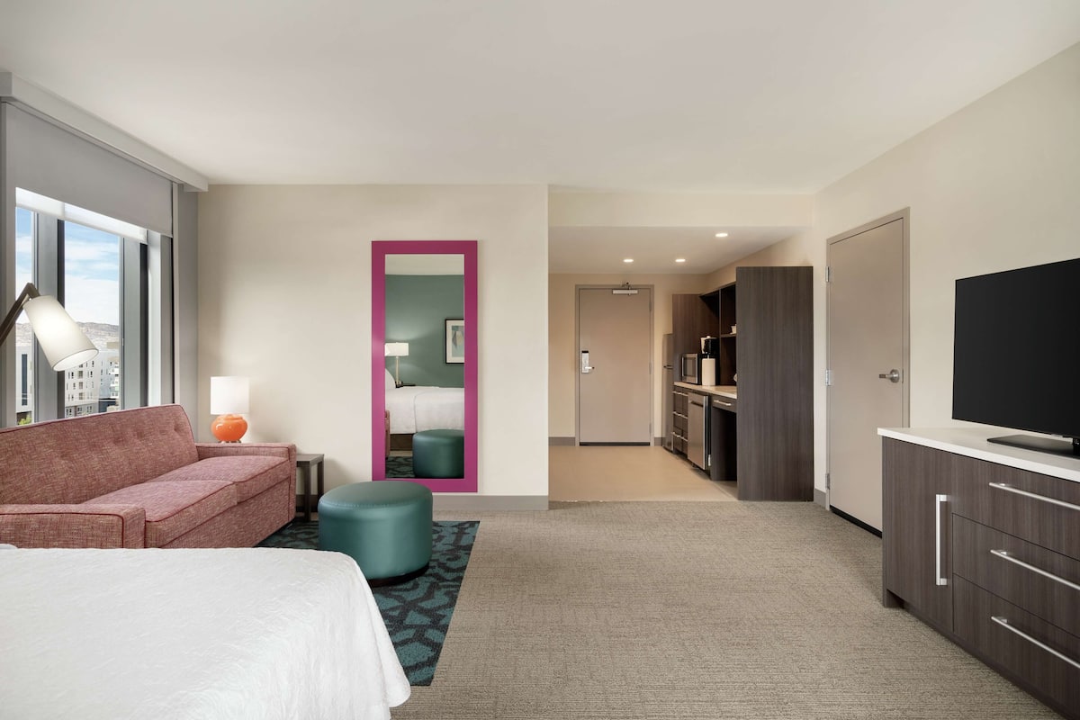 Habitación de hotel con cama, sofá, televisor, escritorio y baño visible a través de una puerta.