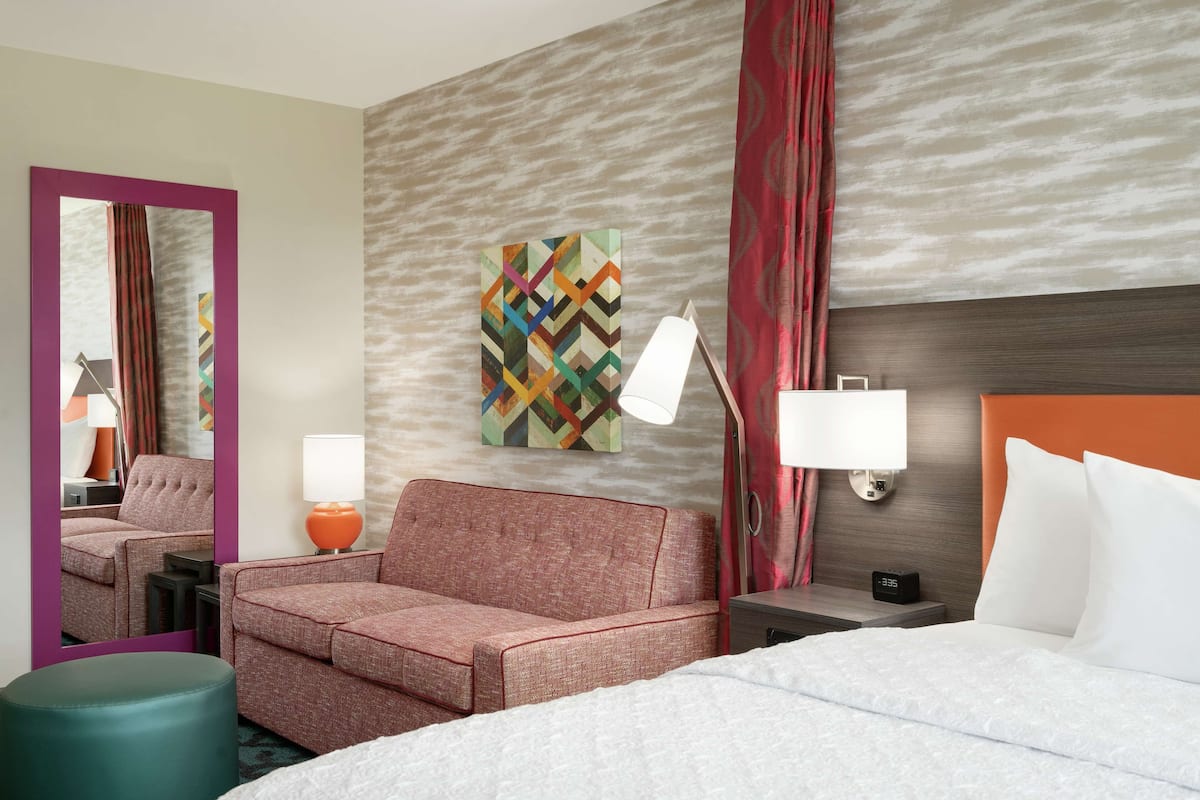 Habitación de hotel con cama, sofá, mesita de noche, lámpara, espejo y pared con una obra de arte geométrica.