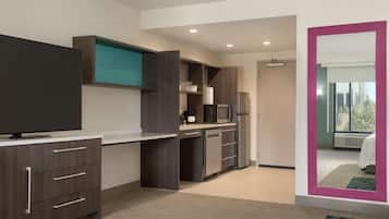 A modern hotel room with a flat-screen TV, a kitchenette, a large mirror, and a view of the outdoors.