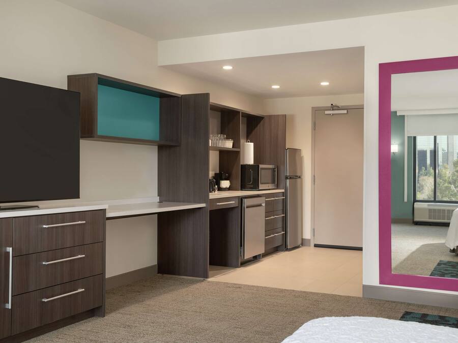 A modern hotel room with a flat-screen TV, a kitchenette, a large mirror, and a view of the outdoors.