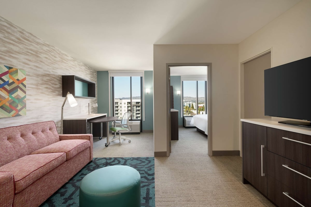 Habitación de hotel con sofá rosa, reposapiés turquesa, escritorio con lámpara, televisor en un mueble y un amplio ventanal con vistas a la ciudad.