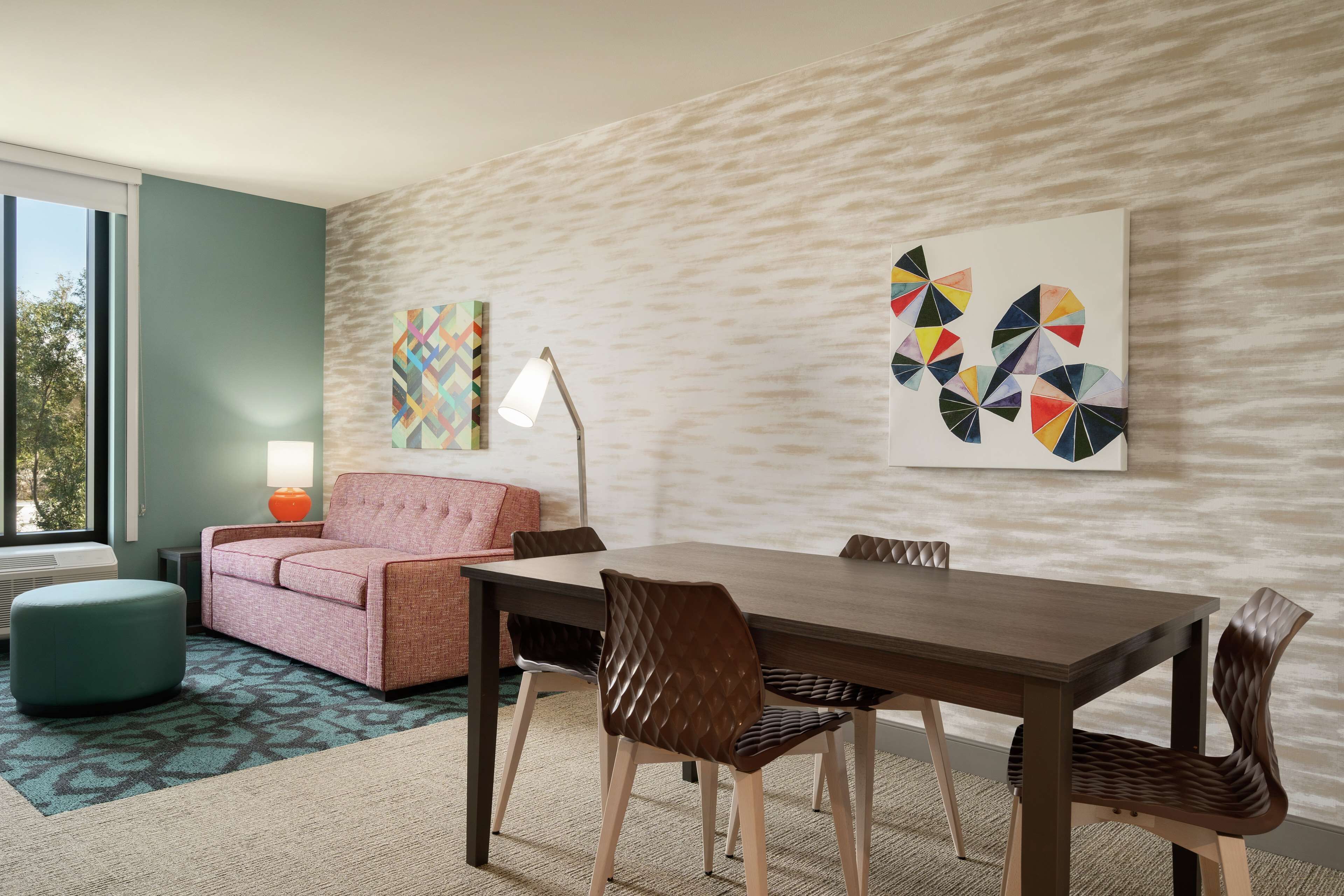 Una habitación de hotel moderna con comedor, un sofá rosa, un reposapiés color turquesa y arte abstracto en la pared.