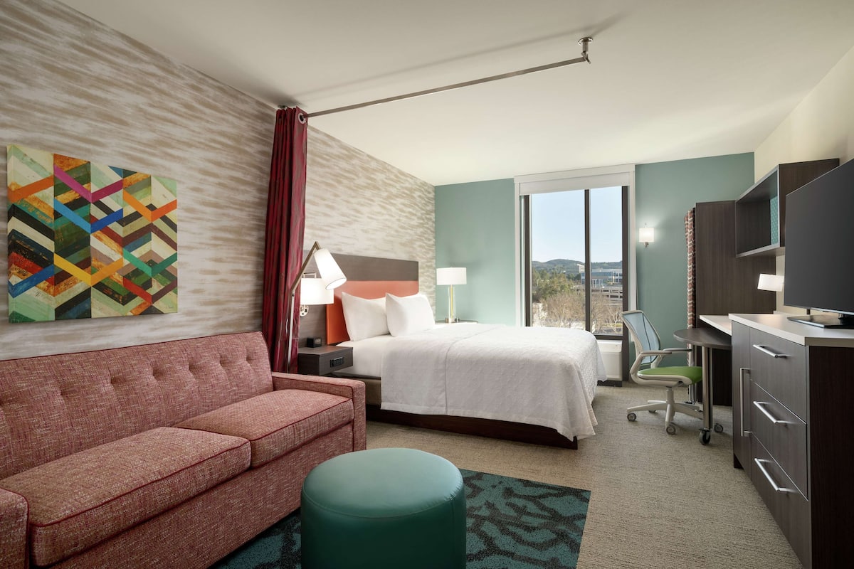 Habitación de hotel con cama, sofá, escritorio con computadora, televisor y ventana con vista al exterior.
