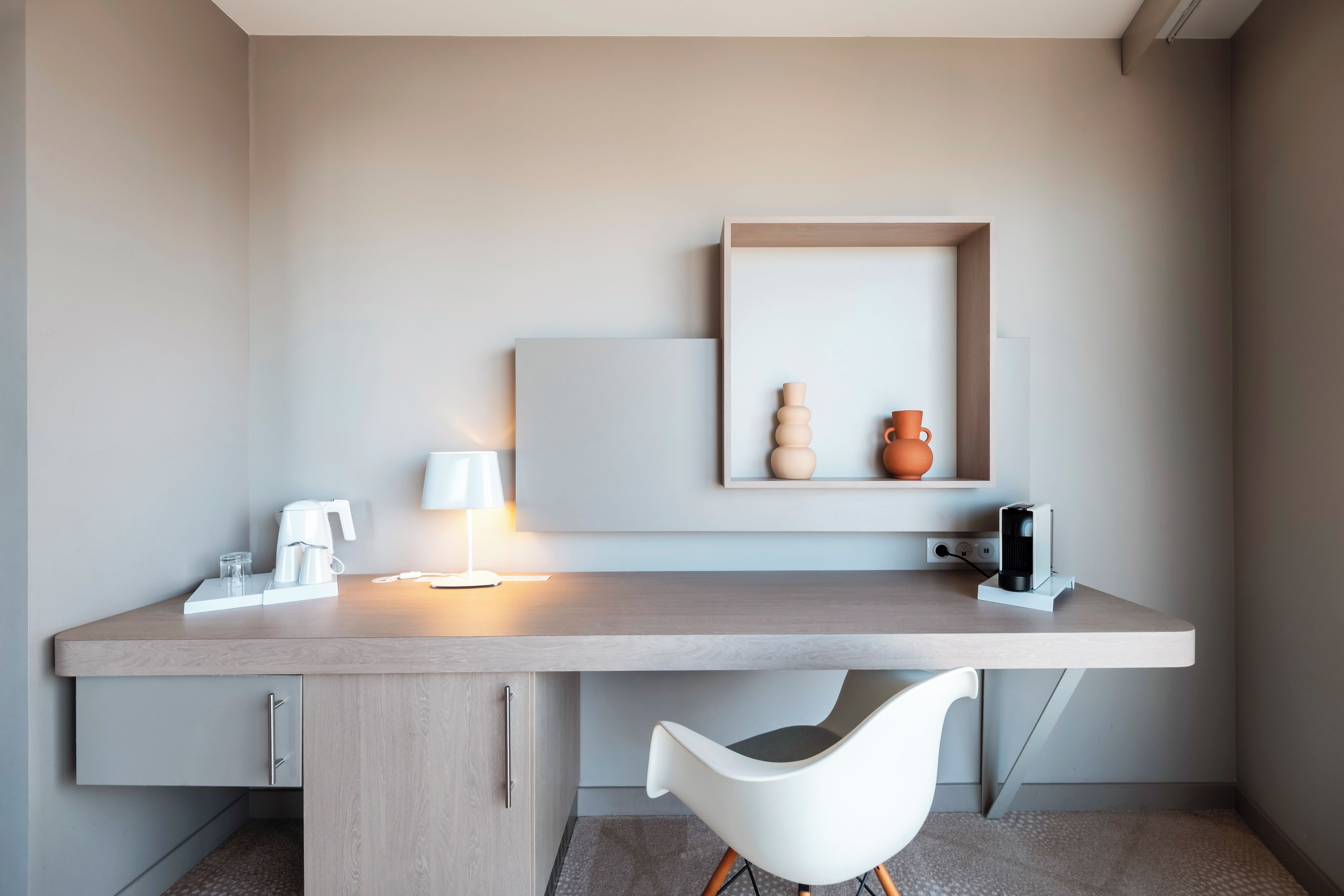 A modern, minimalist workspace with a desk, chair, coffee maker, lamp, and decorative items.
