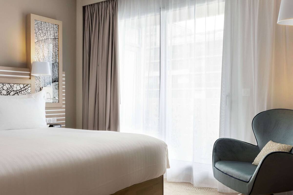 Une chambre d’hôtel moderne avec un grand lit, une chaise et une fenêtre ornée de rideaux légers.