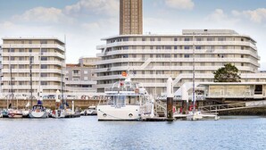 Une vue sur la ville le long du front de mer, avec une tour imposante, un grand bâtiment moderne et plusieurs bateaux amarrés dans une marina.