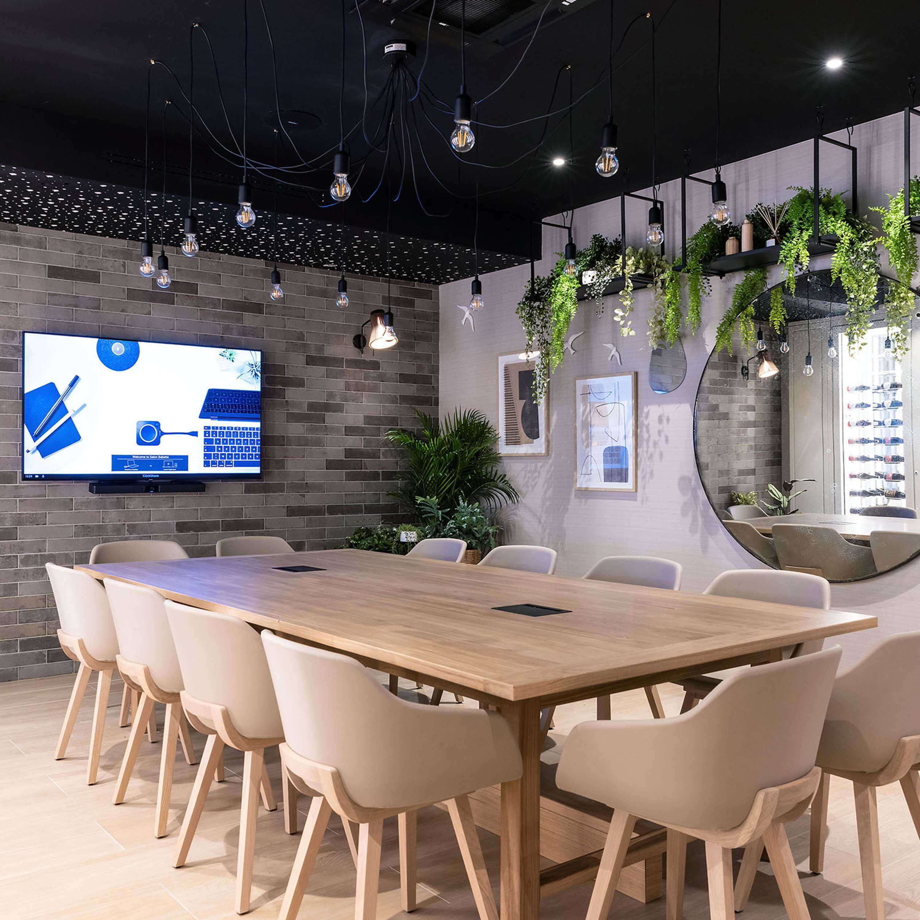 A modern conference room with a long wooden table, beige chairs, a mounted TV on the wall, and hanging plants.