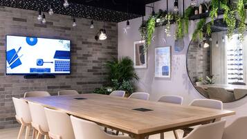 A modern conference room with a long wooden table, beige chairs, a mounted TV on the wall, and hanging plants.