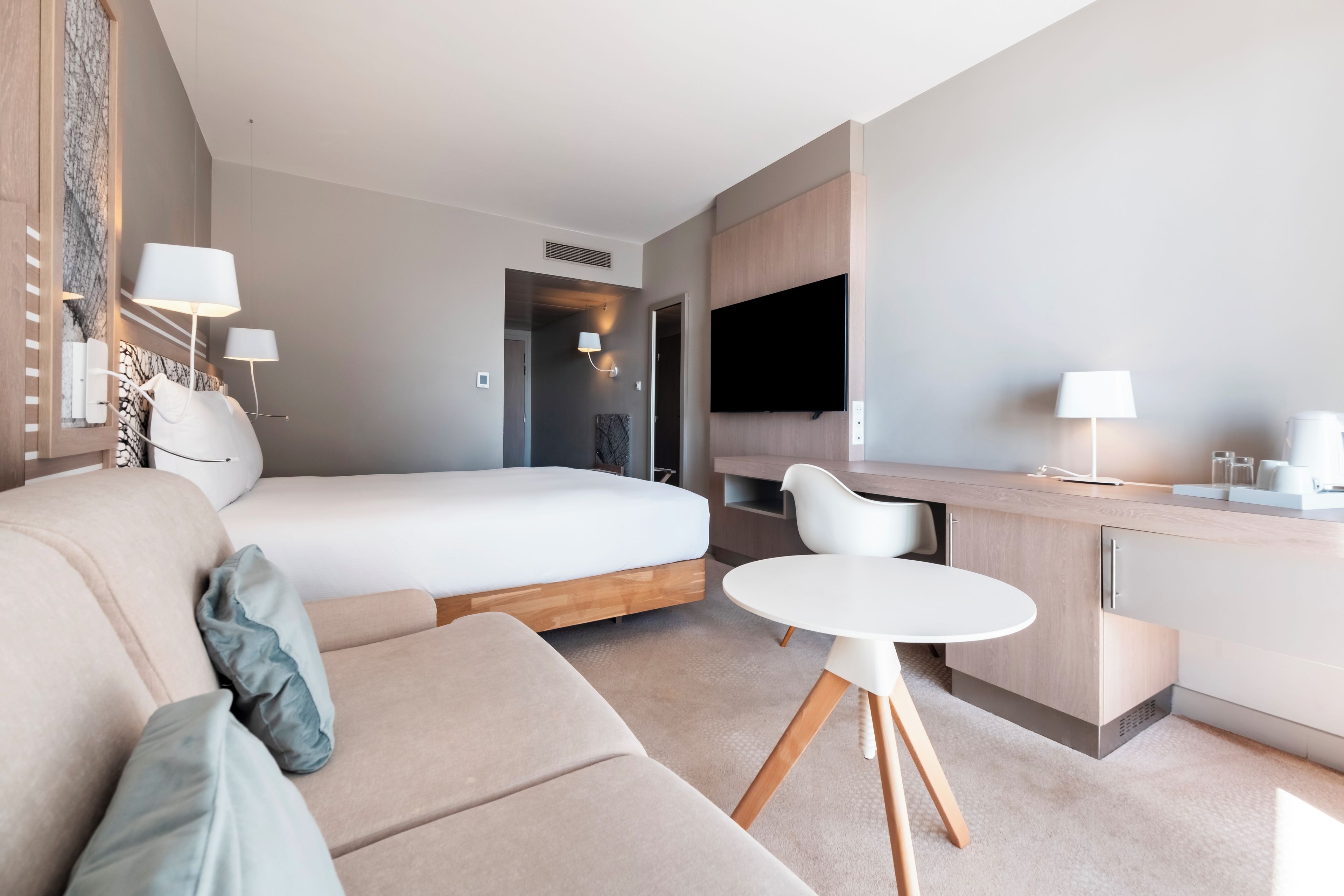 A modern hotel room with a bed, a sofa, a dining table, and a TV.