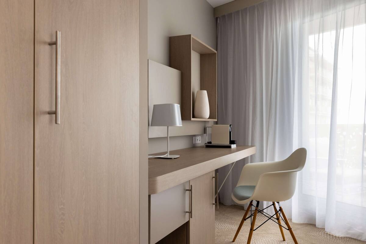 Une chambre moderne avec un bureau en bois, une lampe, une chaise et une fenêtre ornée de rideaux légers.