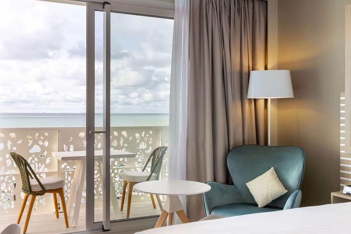 Une chambre d’hôtel avec un lit, une chaise, une table et une vue sur l’océan par une grande fenêtre.