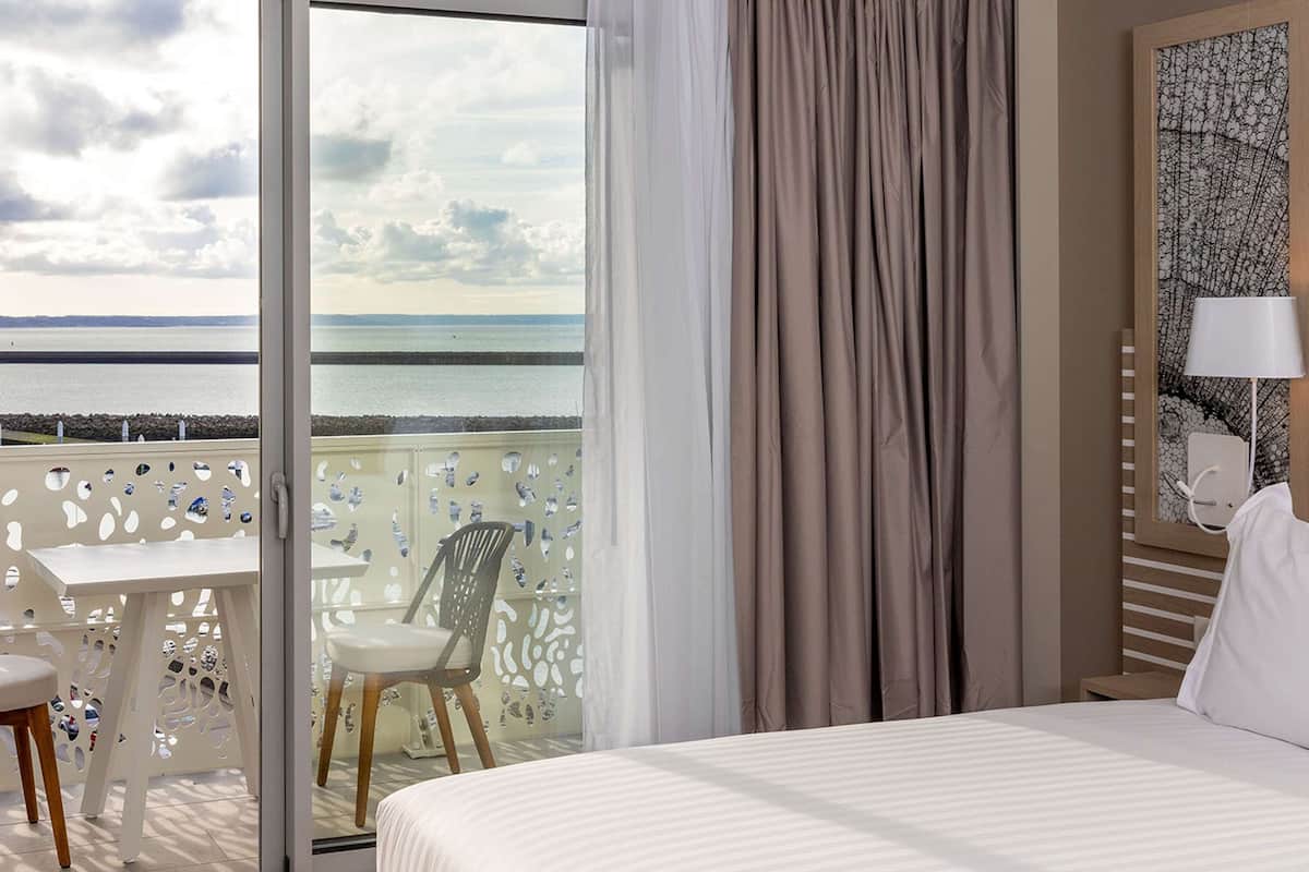 Une chambre d’hôtel avec un lit, une vue sur la mer, un balcon avec une table et des chaises, et une lampe.