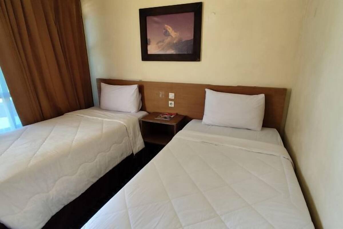 Habitación de hotel con dos camas individuales, una mesita de noche, un cuadro enmarcado en la pared y una ventana con cortinas.
