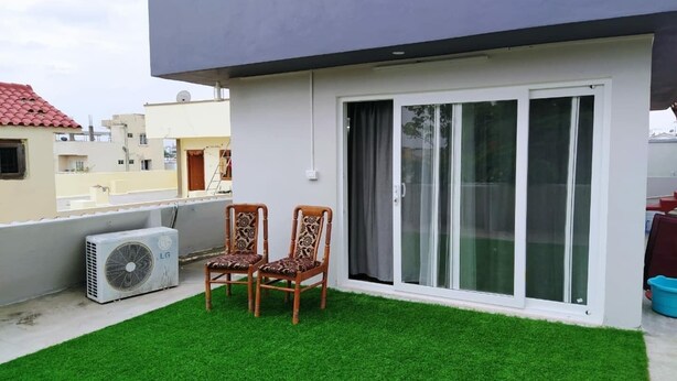 Een modern gebouw met een plat dak, een klein kunstgrasveldje, twee houten stoelen en een grote airconditioner.