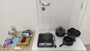 Een kleine kitchenette met een magnetron, koekenpannen, een pan, een gezichtsmasker, een spuitfles en een draagbare speaker op een witte tafel.