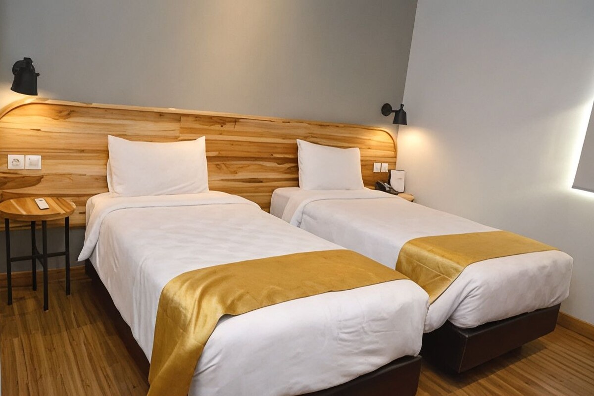 Une chambre d’hôtel avec deux lits, une tête de lit en bois, des lampes de chevet et un petit tabouret.