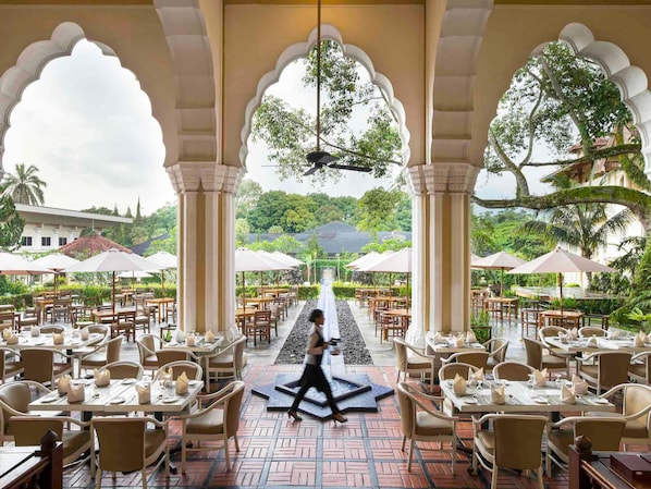 Un espace extérieur aménagé pour les repas, avec des tables et des chaises, une fontaine ou un autre élément aquatique central, et une vue sur un bâtiment traditionnel en arrière-plan.