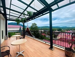 A balcony with a wooden floor, a table, chairs, and a view of a mountainous landscape. - Grand Gallery Bukittinggi (Bukittinggi)