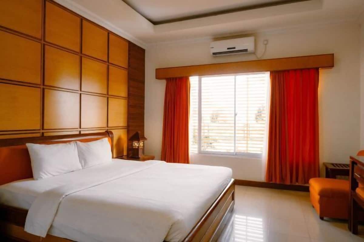 A hotel room with a large bed, a wooden headboard, a nightstand with a lamp, a small table, and a window with red curtains.