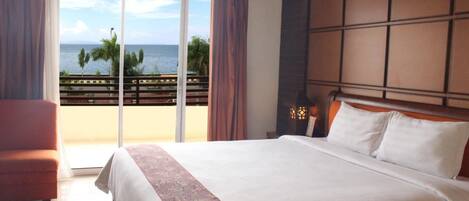 A hotel room with a large bed, a chair, a view of the ocean, and a balcony with a railing.