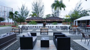 An outdoor seating area with tables and chairs, surrounded by palm trees and a building with a thatched roof. - Sutan Raja Hotel Kolaka (Kolaka)