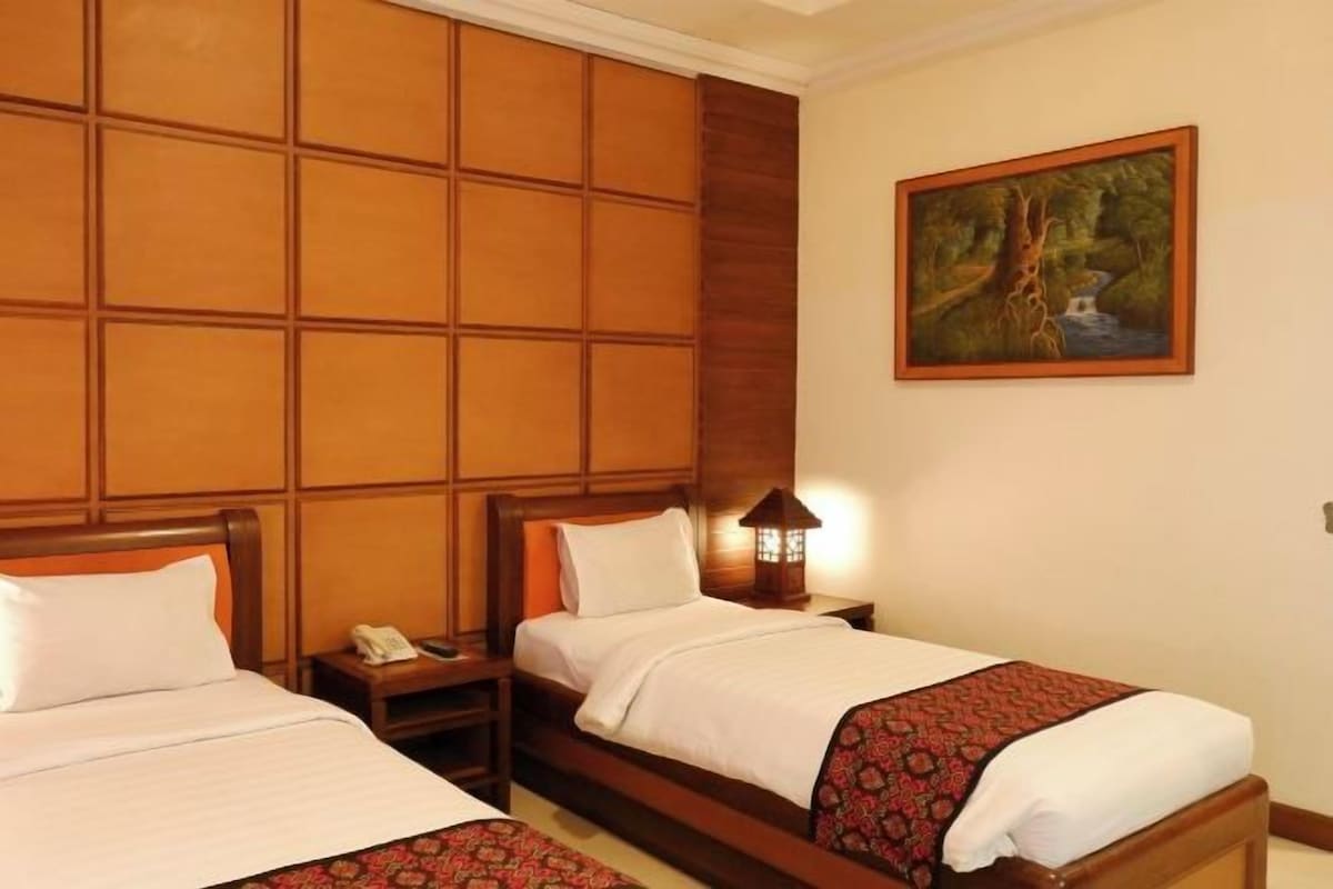 Habitación de hotel con dos camas, cabecera de madera, mesita de noche con lámpara y un cuadro en la pared.