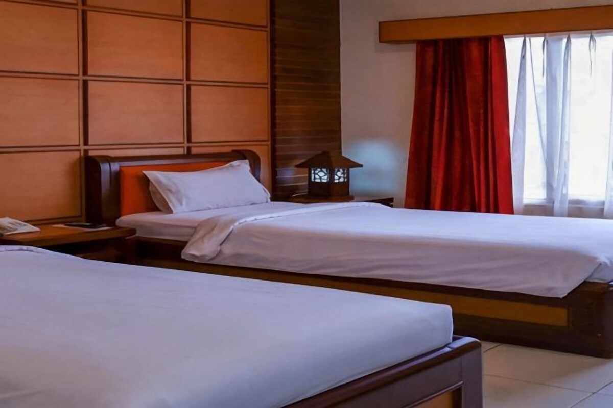 A hotel room with two single beds, a nightstand with a lamp, and a window with red curtains.