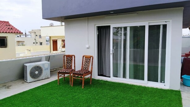 A balcony with artificial grass, two wooden chairs, an air conditioning unit, and a sliding glass door.