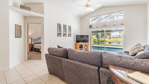 A living room with a large gray sectional sofa, a flat-screen TV on a wooden stand, and a view of a swimming pool through large glass doors. - Turtle Lake - Inspiring Lake Views with Heated Pool by The Bay & Key Collection (Bradenton)