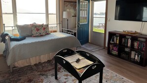 A bedroom with a bed, a chair, a bookshelf, a TV, and a door leading to a balcony with a view of the water. - Sunset Cottage on Pillar Point (Dexter)