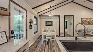 Une cuisine avec un plancher en bois, des meubles de rangement blancs, une table à manger avec des chaises, un luminaire suspendu et une grande fenêtre.