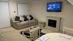 A living room with a beige sofa, a fireplace, a television mounted on the wall, and a dining table with a marble placemat. - Cheerful 3 Bedroomed house,with parking and free intern free (Luton)