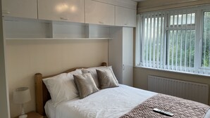 A bedroom with a bed, bedside table, lamp, and a window with blinds. - Cheerful 3 Bedroomed house,with parking and free intern free (Luton)