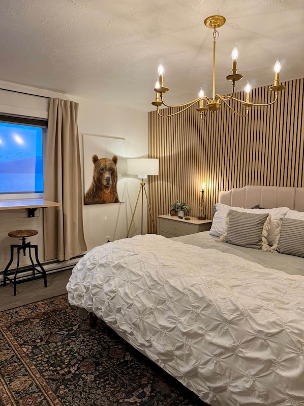 A bedroom with a large bed, a desk, a bear wall art, a chandelier, and a view of the mountains through the window.