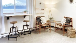 Una habitación acogedora con vistas a montañas, una silla de madera con cojines, una mesita con una lámpara y dos taburetes.