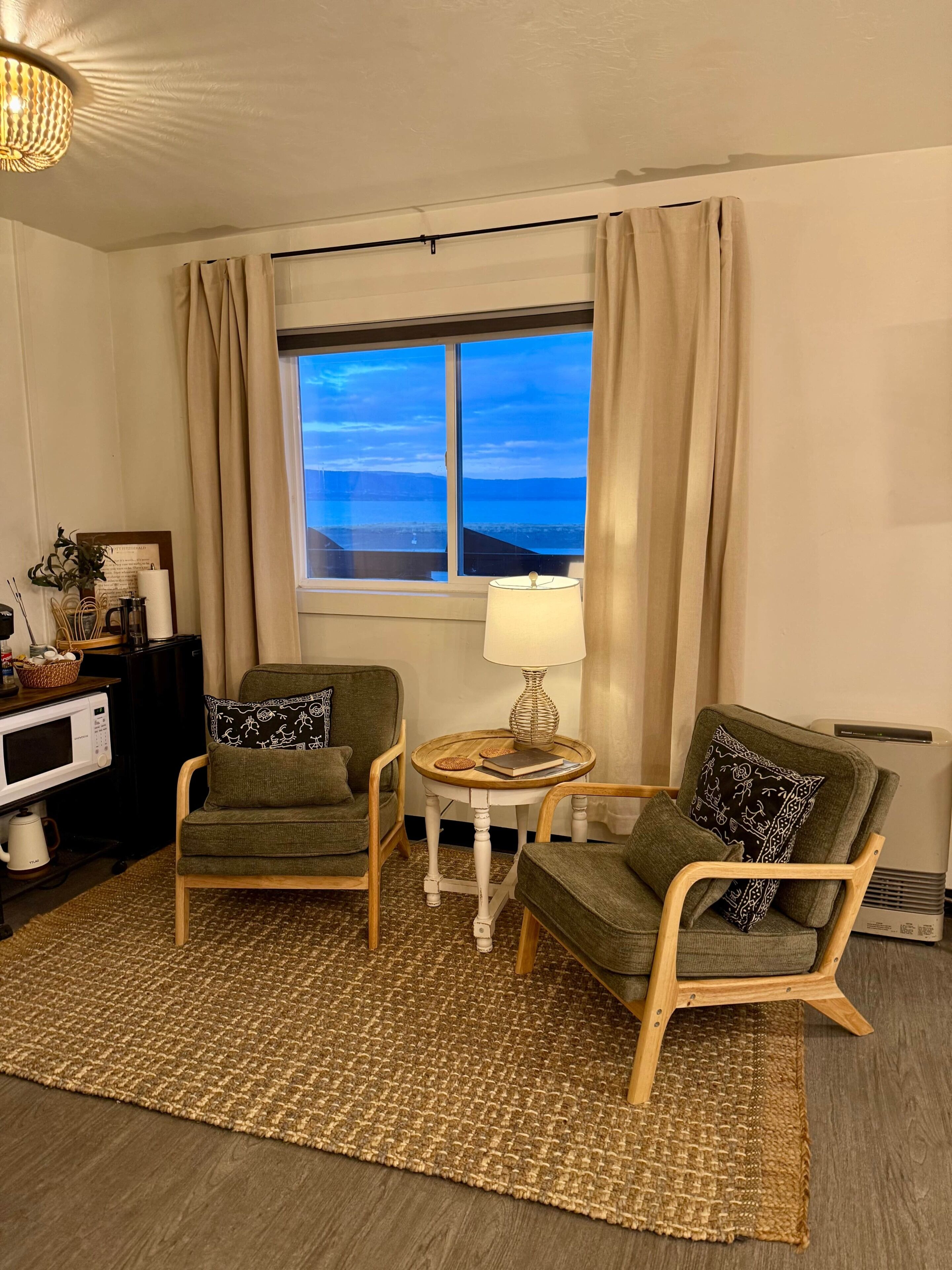 Ein Zimmer mit zwei grünen Sesseln, einem kleinen runden Tisch mit Lampe, einem Fenster mit Vorhängen, einer Mikrowelle und einem Heizkörper.