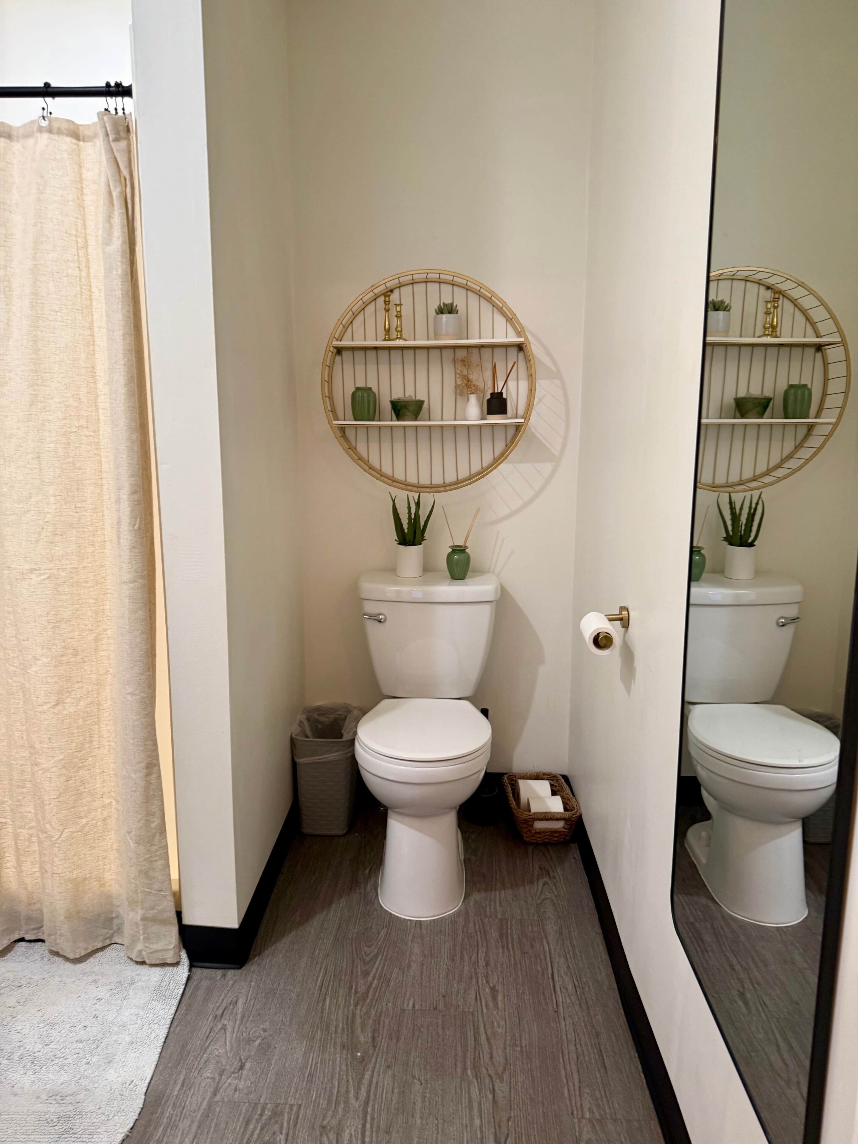 A bathroom with two toilets, a round shelf with plants, a white toilet paper holder, a trash bin, and a wooden floor.