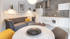 Uma sala de estar moderna com um sofá cinzento, uma mesa redonda de mármore e uma área de cozinha com máquina de lavar.
