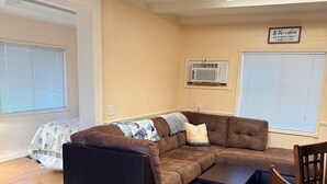 Una sala de estar con un sofá seccional, una mesa de centro y una silla de madera. Hay una ventana con persianas, un equipo de aire acondicionado y un letrero en la pared.