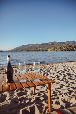Ein Tisch mit einer Flasche und Gläsern an einem sandigen Strand, im Hintergrund ein See und Berge.