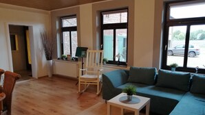 Un salon avec un plancher en bois, un canapé vert, un fauteuil à bascule blanc, une petite table blanche et une fenêtre donnant sur un mur de briques et des voitures garées.